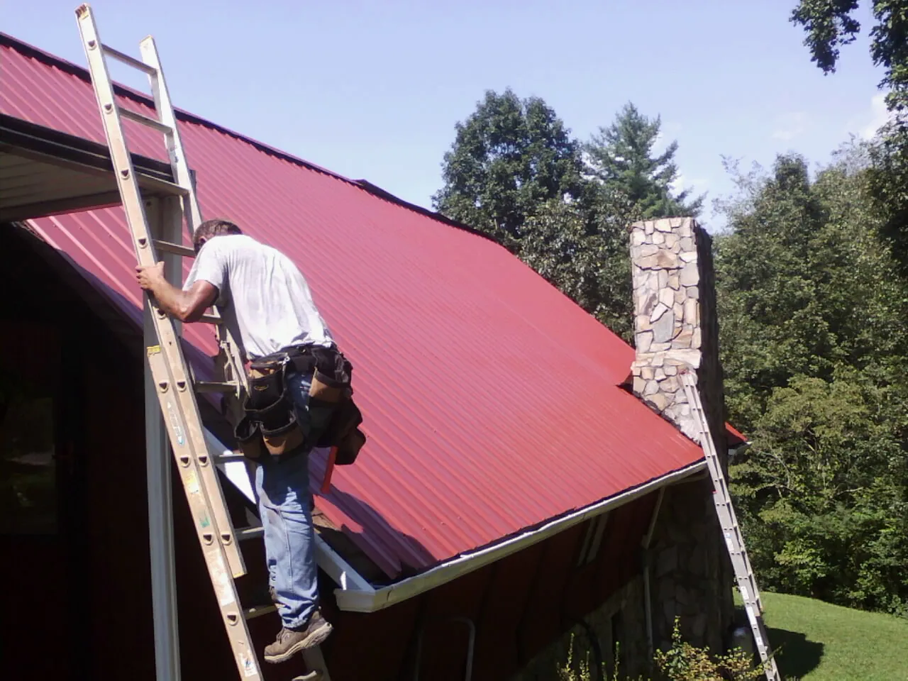 Roofer on a ladder beside a red metal roof and stone chimney — Soffit & Fascia Repair by Roofing Brothers in Oak Grove